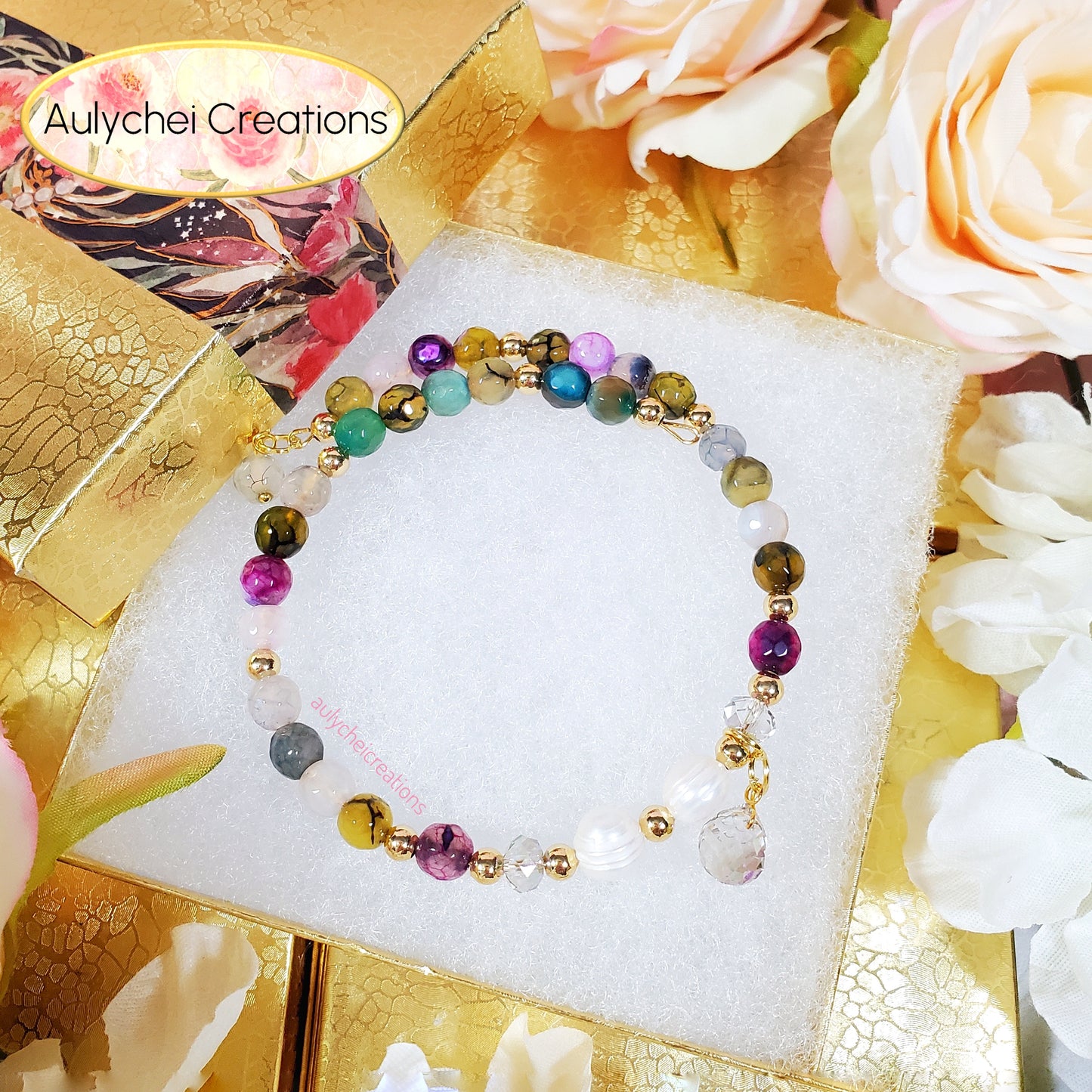 Multicolored beaded bracelet on a white stand with flowers and gold accents in the background, featuring 'Aulychei Creations' branding.
