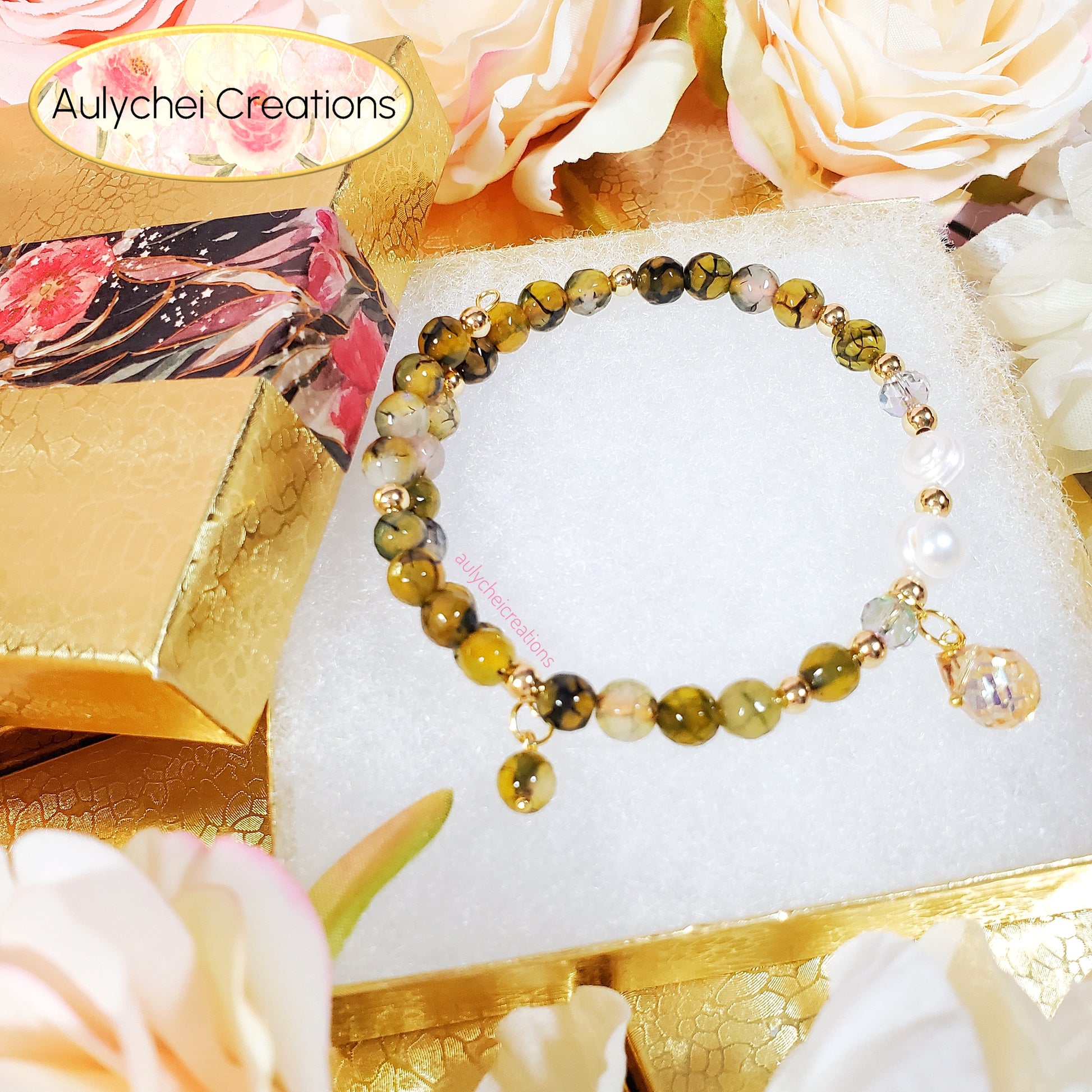 Olive green beaded gemstone bracelet on a white cotton surface, surrounded by flowers and a decorative box, with 'Aulychei Creations' branding.